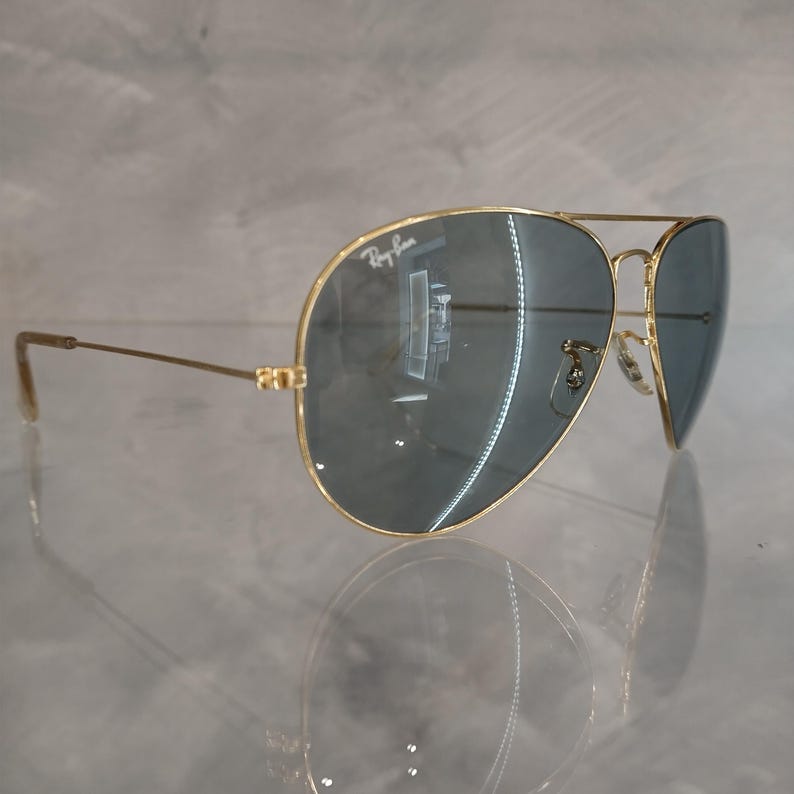 Puede incluir: Gafas de sol aviador doradas con lentes grises oscuros. El logotipo de Ray-Ban es visible en la lente izquierda.