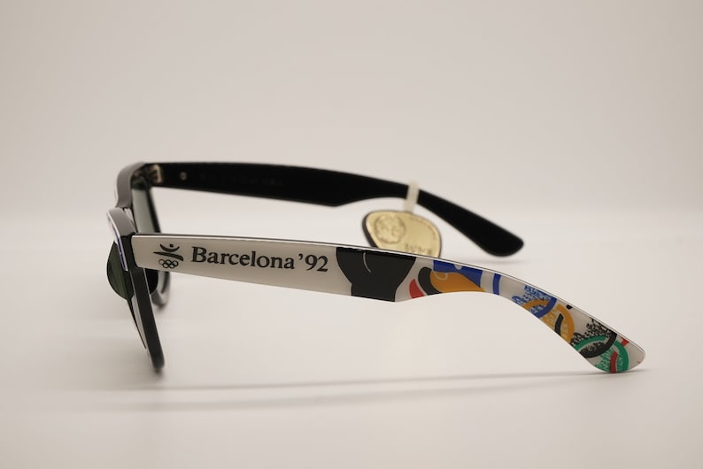 K&ouml;nnte beinhalten: Schwarze Sonnenbrille mit silbernem Rahmen und einem bunten olympischen Design. Der Rahmen tr&auml;gt den Schriftzug "Barcelona '92".
