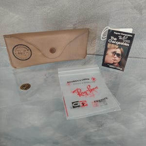 May include: A brown leather case for Ray-Ban sunglasses with a Ray-Ban logo and a small tag that says "The Changeables" with a picture of a couple wearing sunglasses.