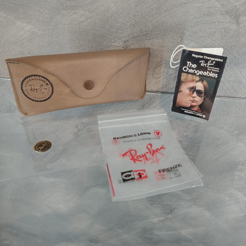 May include: A brown leather case for Ray-Ban sunglasses with a Ray-Ban logo and a small tag that says "The Changeables" with a picture of a couple wearing sunglasses.