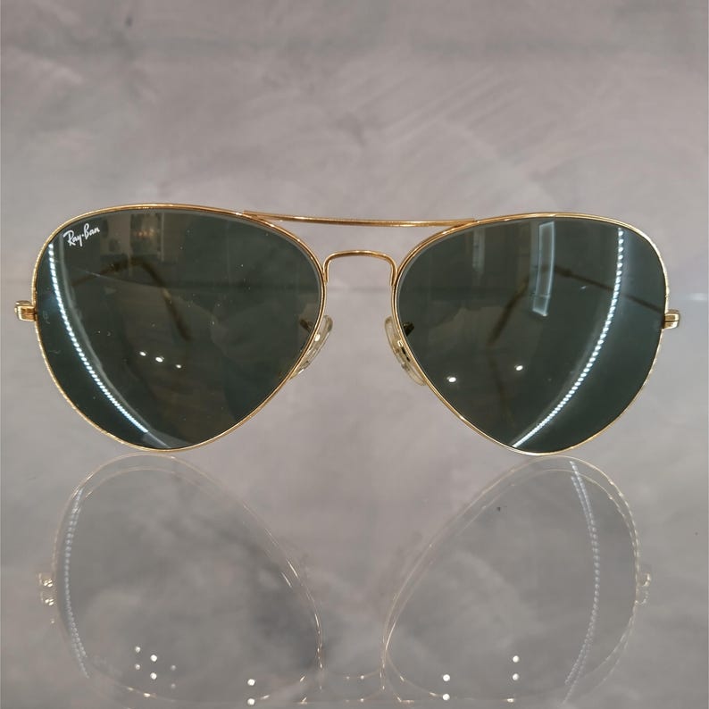 Puede incluir: Gafas de sol aviador doradas con lentes verde oscuro. Las gafas tienen el logotipo de Ray-Ban en la lente izquierda.