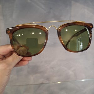 May include: Brown sunglasses with a gold-colored metal bridge and green lenses. The sunglasses have a classic, rectangular shape with a double bridge design. The frames are a translucent brown color.