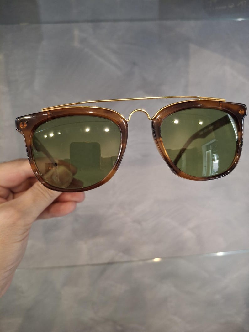 May include: Brown sunglasses with a gold-colored metal bridge and green lenses. The sunglasses have a classic, rectangular shape with a double bridge design. The frames are a translucent brown color.