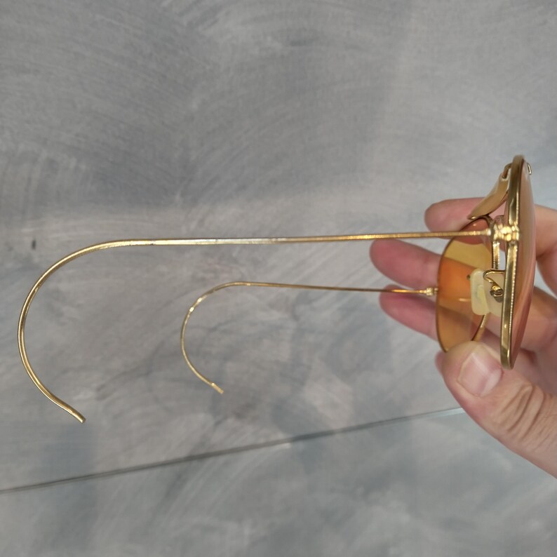 May include: Gold-framed sunglasses with orange-tinted lenses. The sunglasses have a classic aviator shape with a double bridge and curved temples. The lenses are a warm, amber color, and the frames are a shiny gold.
