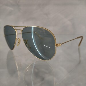 May include: Gold-framed aviator sunglasses with blue-tinted lenses. The sunglasses have a classic teardrop shape and a double bridge. The temples are thin and gold, with a slight curve at the ends. The lenses reflect light.