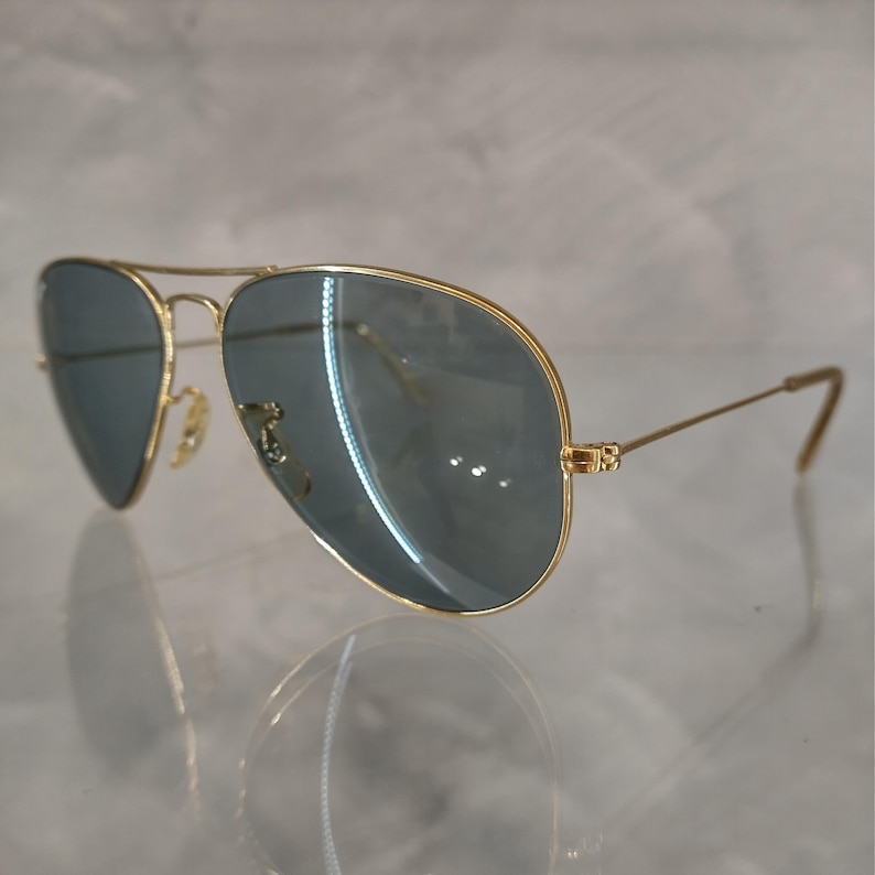 May include: Gold-framed aviator sunglasses with blue-tinted lenses. The sunglasses have a classic teardrop shape and a double bridge. The temples are thin and gold, with a slight curve at the ends. The lenses reflect light.