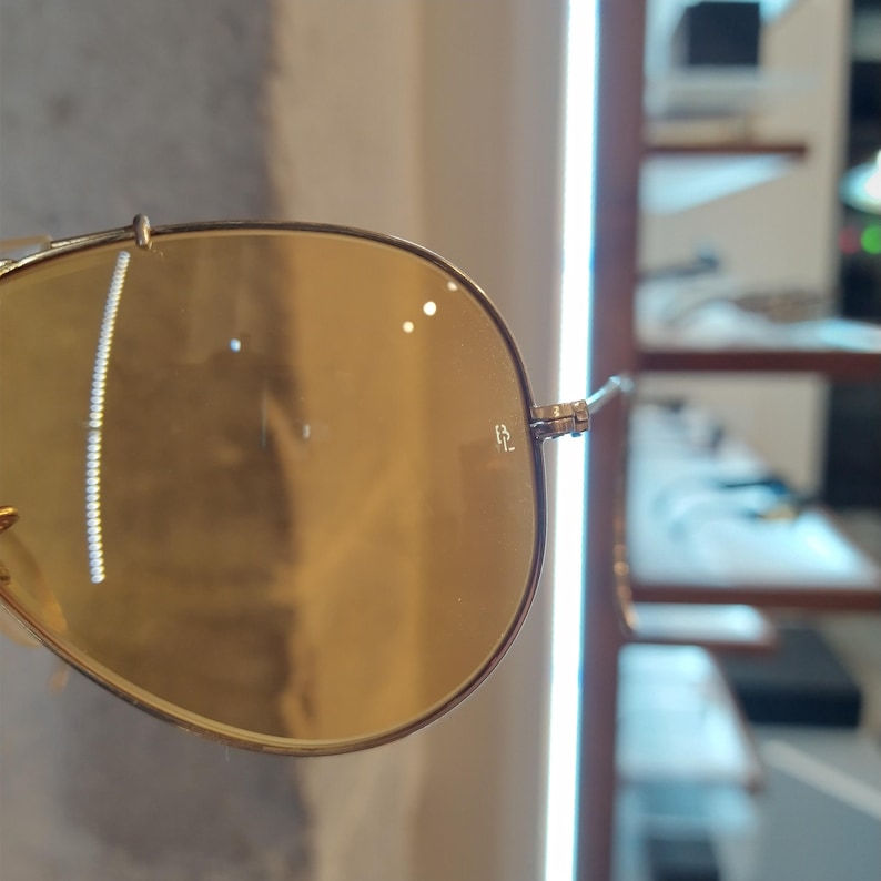 May include: Close-up of gold-rimmed sunglasses with amber-tinted lenses. The glasses have a classic aviator style with a small logo etched on the lens. The frame is a light gold color.