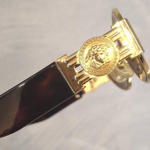 K&ouml;nnte beinhalten: Nahaufnahme einer goldfarbenen Sonnenbrille mit einem Medusa-Kopf-Emblem am B&uuml;gel. Der Rahmen hat ein dunkelbraunes Schildpattmuster. Die Sonnenbrille hat ein schlankes, modernes Design.