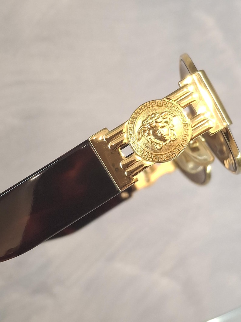 K&ouml;nnte beinhalten: Nahaufnahme einer goldfarbenen Sonnenbrille mit einem Medusa-Kopf-Emblem am B&uuml;gel. Der Rahmen hat ein dunkelbraunes Schildpattmuster. Die Sonnenbrille hat ein schlankes, modernes Design.