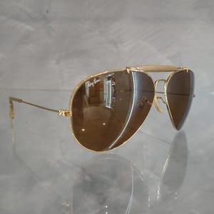 May include: Gold aviator sunglasses with brown lenses. The sunglasses have a metal frame and a bridge with a gold bar.