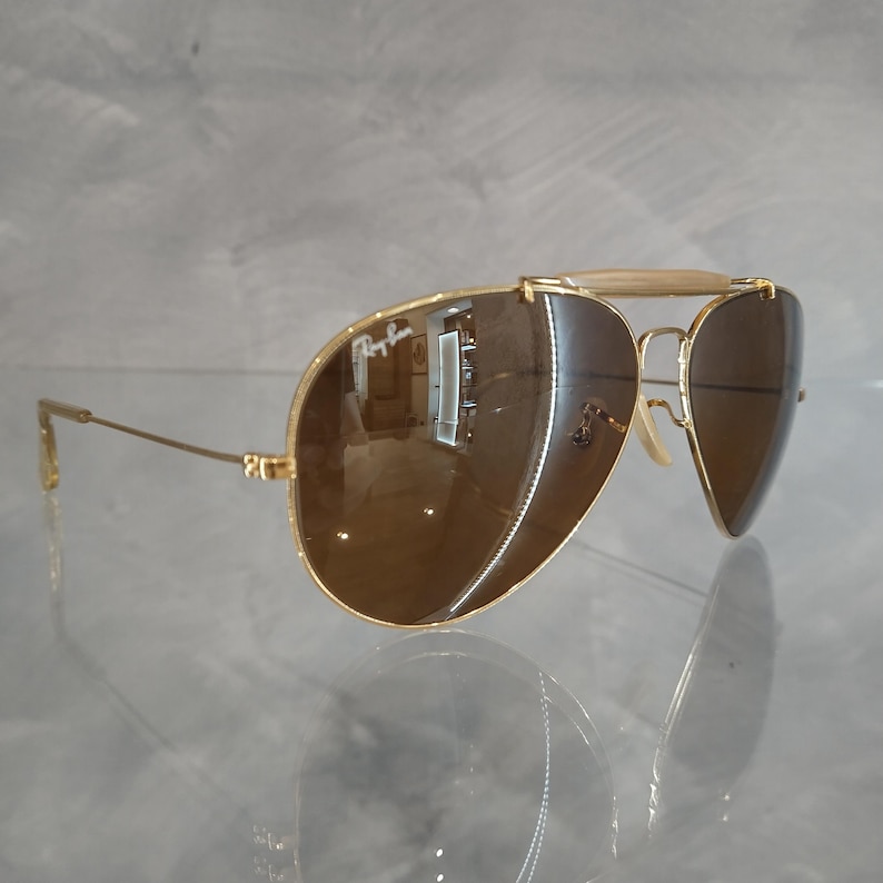 May include: Gold aviator sunglasses with brown lenses. The sunglasses have a metal frame and a bridge with a gold bar.