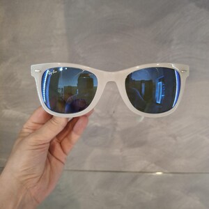 Pu&ograve; includere: Occhiali da sole bianchi con lenti blu riflettenti. Gli occhiali hanno un classico design a montatura rettangolare. Il logo Ray-Ban &egrave; visibile sulla lente sinistra. Gli occhiali sono tenuti su uno sfondo grigio.