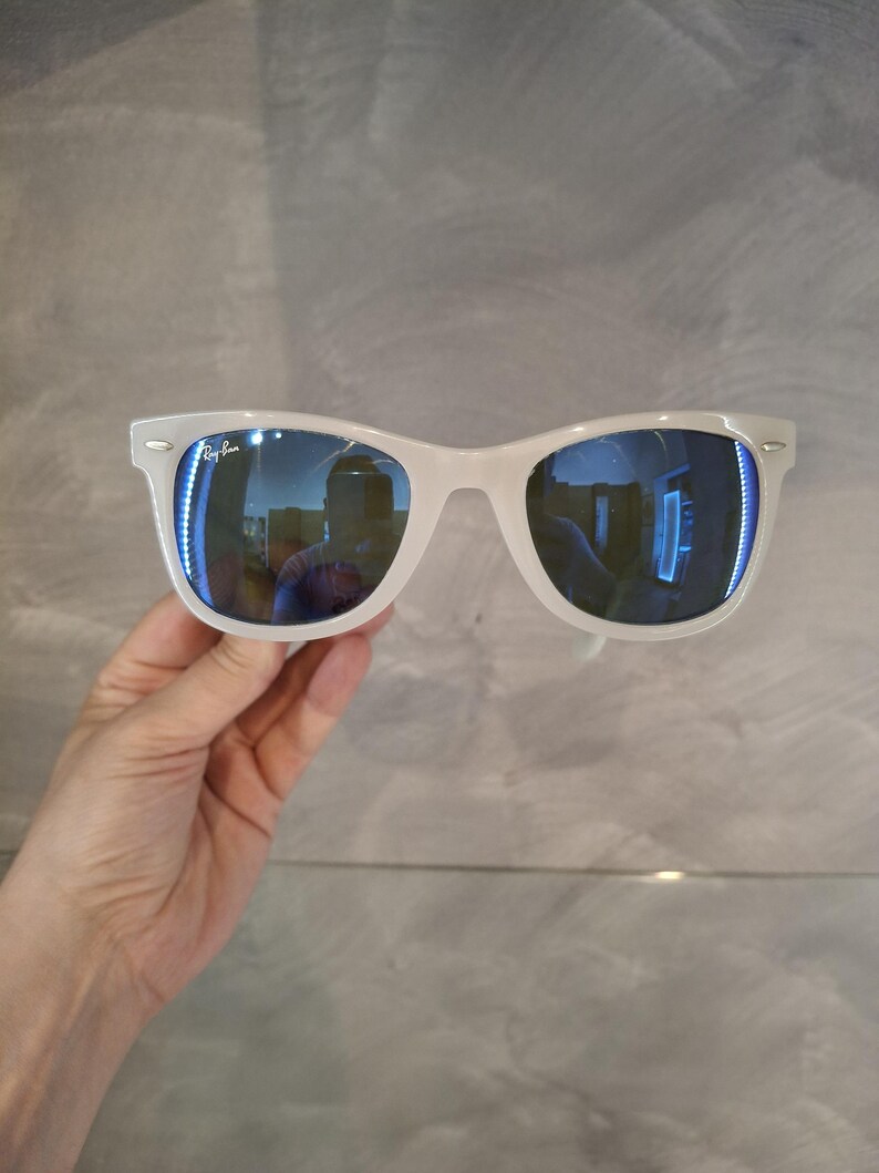 Pu&ograve; includere: Occhiali da sole bianchi con lenti blu riflettenti. Gli occhiali hanno un classico design a montatura rettangolare. Il logo Ray-Ban &egrave; visibile sulla lente sinistra. Gli occhiali sono tenuti su uno sfondo grigio.