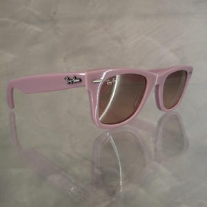 Puede incluir: Gafas de sol rosas con lentes marrones. Las gafas de sol tienen un dise&ntilde;o de montura rectangular cl&aacute;sico. El logotipo de Ray-Ban es visible en la patilla. Las gafas de sol se reflejan en la superficie brillante de abajo.