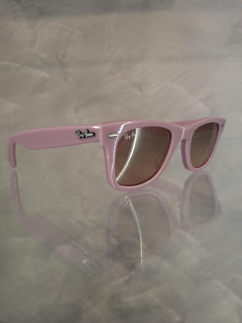 Puede incluir: Gafas de sol rosas con lentes marrones. Las gafas de sol tienen un dise&ntilde;o de montura rectangular cl&aacute;sico. El logotipo de Ray-Ban es visible en la patilla. Las gafas de sol se reflejan en la superficie brillante de abajo.
