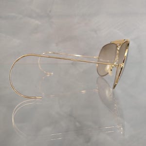 May include: Gold aviator sunglasses with brown gradient lenses and a thin metal frame. The sunglasses have a wire temple arm that curves around the ear.
