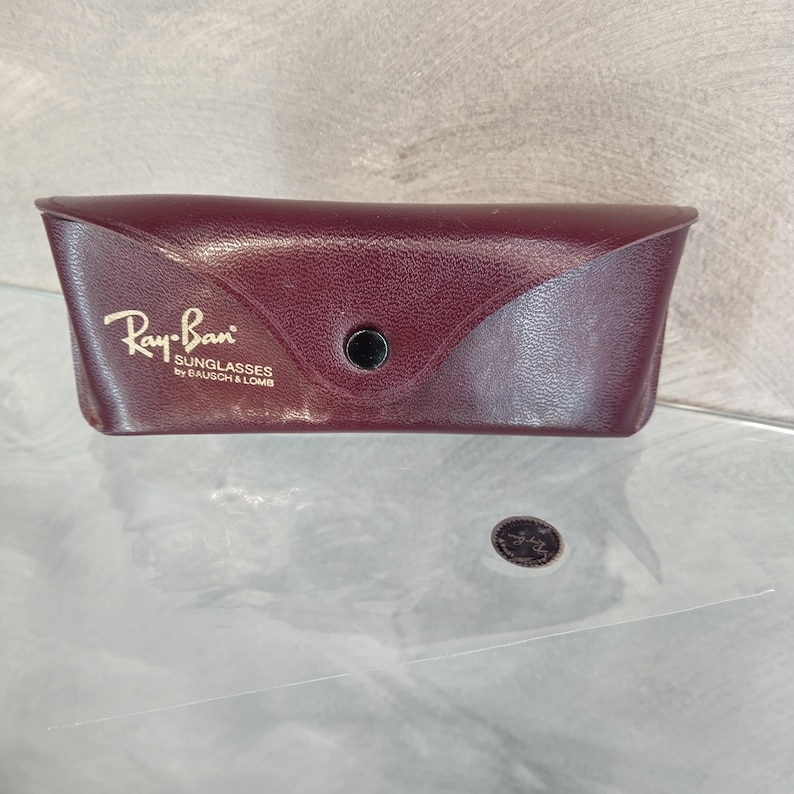 Op de afbeelding: Een bordeauxrode leren etui voor Ray-Ban zonnebrillen. De etui heeft een goudkleurig reli&euml;flogo met de tekst "Ray-Ban Sunglasses by Bausch & Lomb".