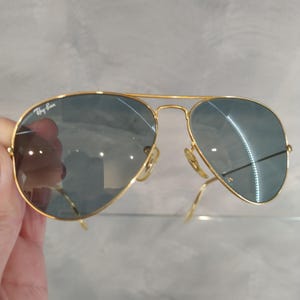 May include: Gold-framed aviator sunglasses with blue-tinted lenses. The Ray-Ban logo is visible on the upper left lens. The sunglasses have a double bridge and curved temples.
