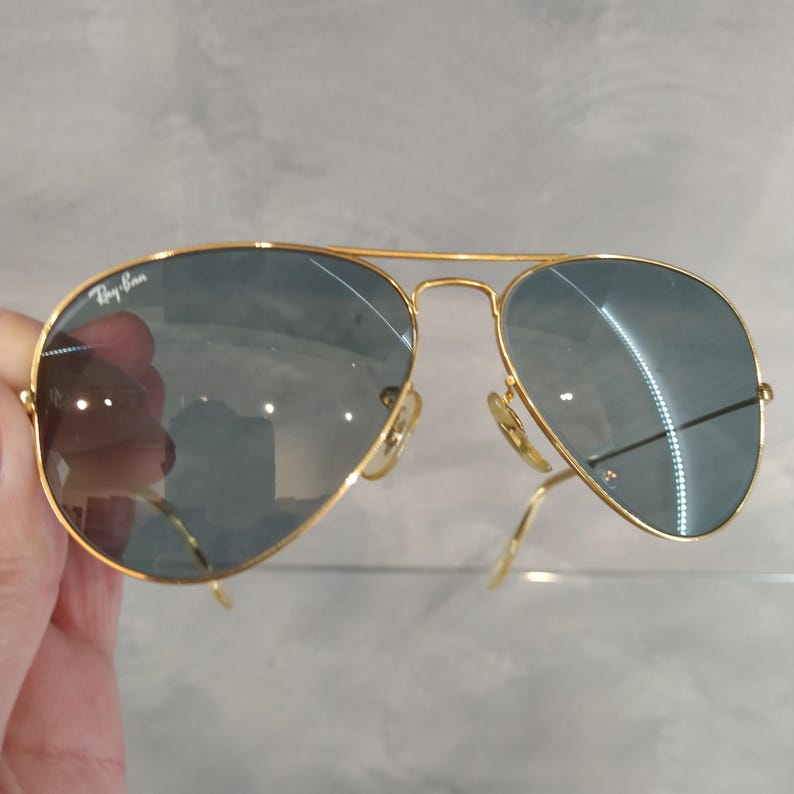 May include: Gold-framed aviator sunglasses with blue-tinted lenses. The Ray-Ban logo is visible on the upper left lens. The sunglasses have a double bridge and curved temples.