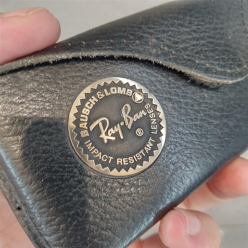 Puede incluir: Una funda de cuero negro para gafas de sol Ray-Ban con un logotipo de metal plateado que dice "Bausch & Lomb Ray-Ban Impact Resistant Lenses".