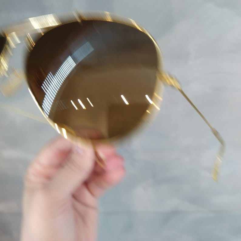 May include: Gold-toned aviator sunglasses with brown lenses. The sunglasses are held in a hand.