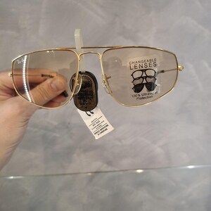 May include: Gold-coloured aviator-style spectacles with clear lenses. The spectacles have a double bridge and a tag that reads "CHANGEABLE LENSES" and "100% Ultraviolet Protection". The lenses are slightly tinted. The spectacles are held by a hand.