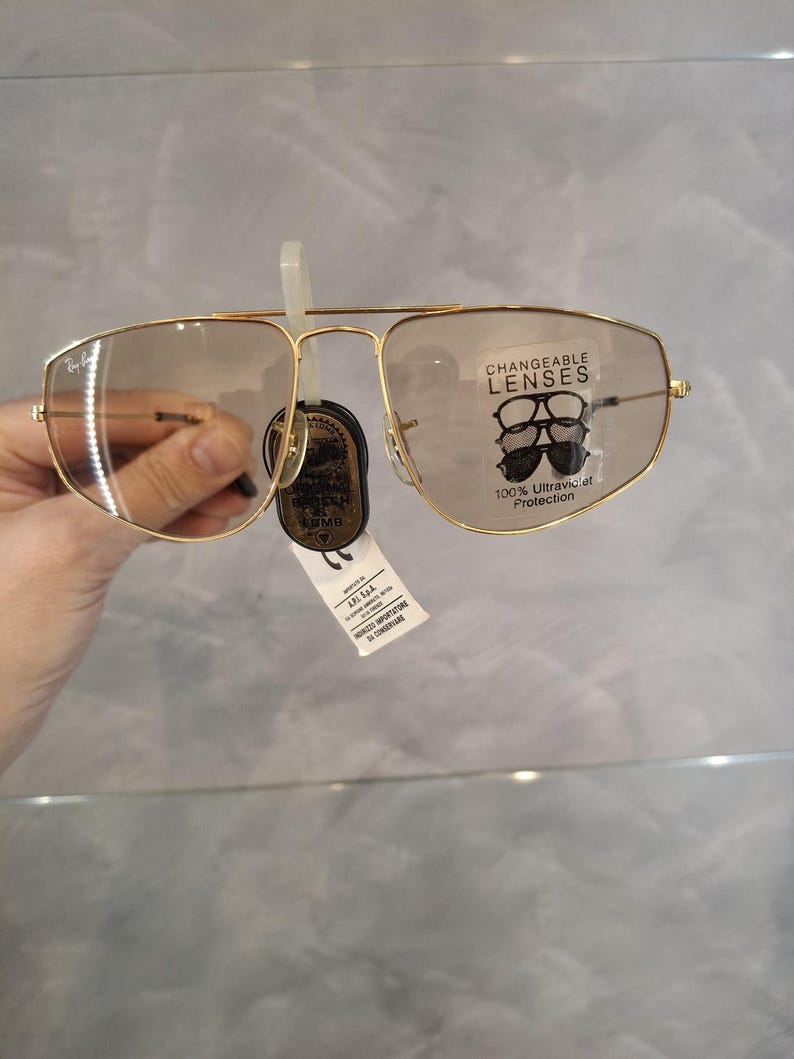 May include: Gold-coloured aviator-style spectacles with clear lenses. The spectacles have a double bridge and a tag that reads "CHANGEABLE LENSES" and "100% Ultraviolet Protection". The lenses are slightly tinted. The spectacles are held by a hand.