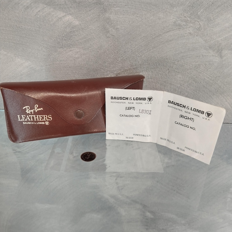 Op de afbeelding: Een bruin leren Ray-Ban etui met een drukknoop. Het etui heeft een label met de tekst "Ray-Ban Leathers" en "Bausch & Lomb". Het etui heeft ook twee witte labels met de tekst "Bausch & Lomb" en het catalogusnummer "L8301".