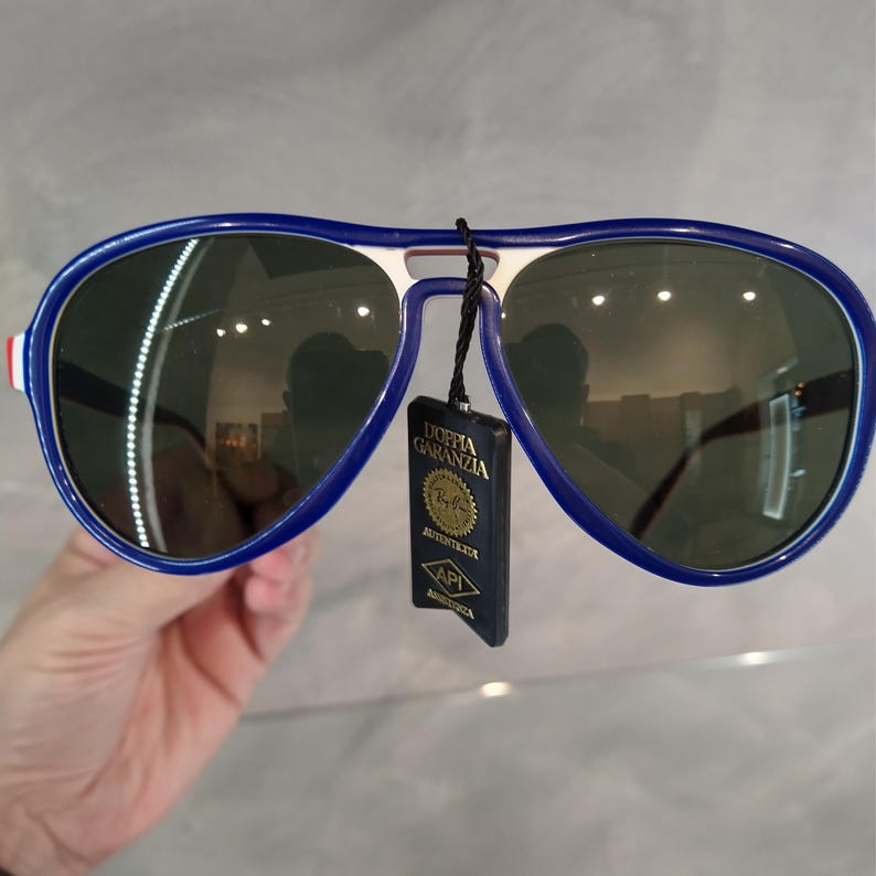 May include: Blue and white aviator sunglasses with green lenses. The sunglasses have a tag attached that says "Doffia Garanzia" and "Autentica Ray-Ban".