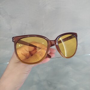 May include: Brown cat-eye sunglasses with yellow lenses. The sunglasses are held in a hand.
