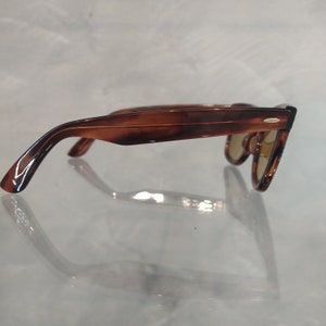 May include: Brown tortoiseshell sunglasses with a gold accent on the frame. The sunglasses are on a white surface.