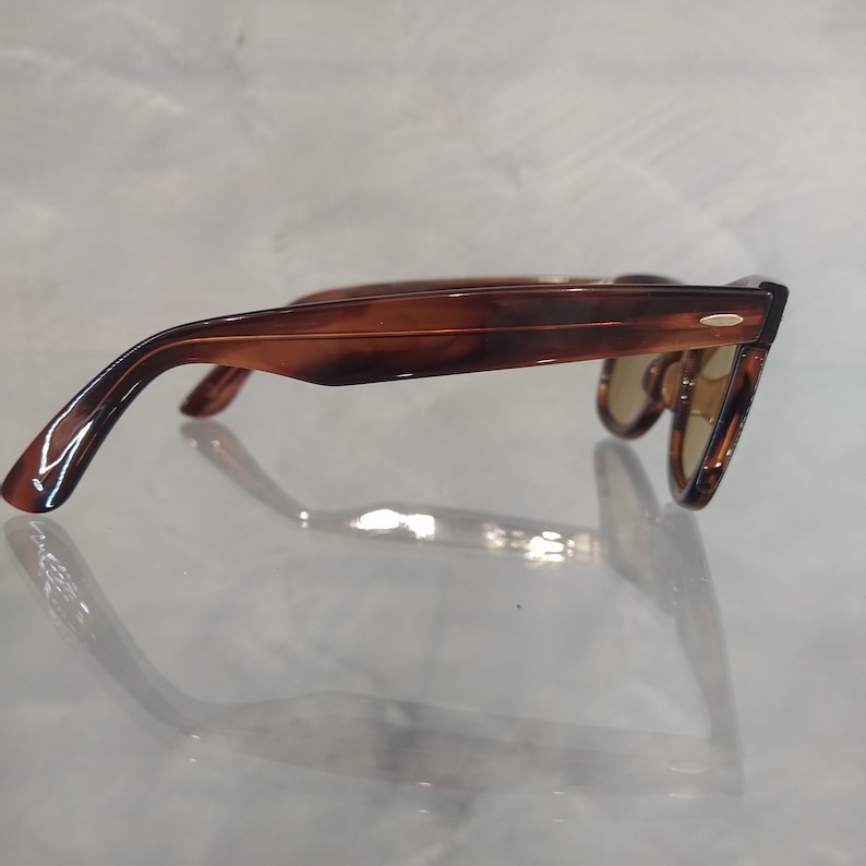 May include: Brown tortoiseshell sunglasses with a gold accent on the frame. The sunglasses are on a white surface.