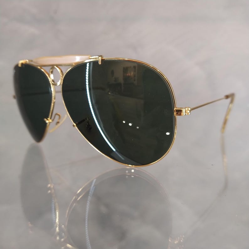 Puede incluir: Gafas de sol aviador doradas con lentes verdes y un puente blanco. Las gafas est&aacute;n sobre una superficie blanca.