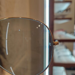 Puede incluir: Primer plano de una gafas de sol aviador con lentes azules. Las gafas tienen un marco dorado y el texto "RL" es visible en la lente derecha.