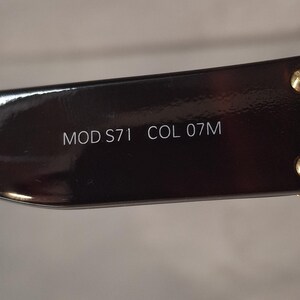 K&ouml;nnte beinhalten: Nahaufnahme eines Teils eines dunkelbraunen Brillengestells. Der Text "MOD S71 COL 07M" ist wei&szlig; auf dem Gestell aufgedruckt. Das Gestell hat eine gl&auml;nzende Oberfl&auml;che und kleine goldfarbene Akzente.