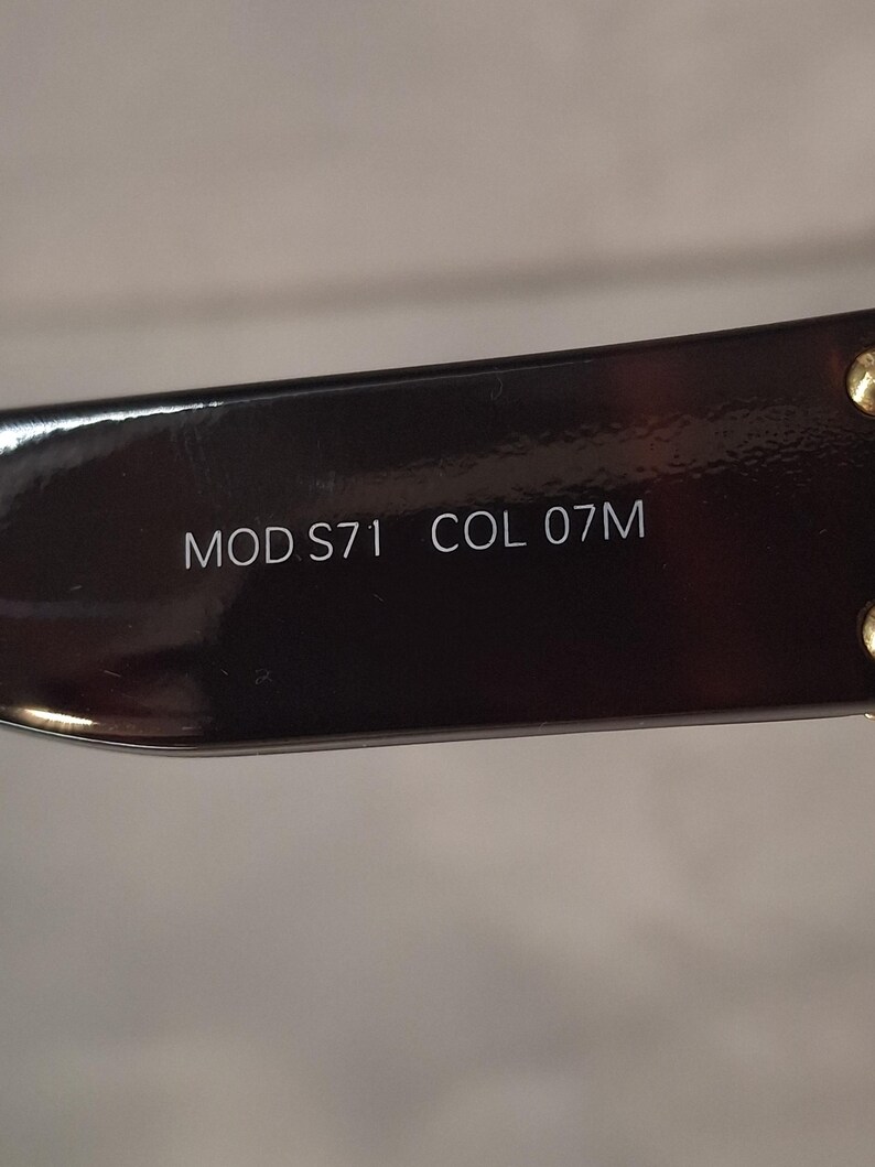 K&ouml;nnte beinhalten: Nahaufnahme eines Teils eines dunkelbraunen Brillengestells. Der Text "MOD S71 COL 07M" ist wei&szlig; auf dem Gestell aufgedruckt. Das Gestell hat eine gl&auml;nzende Oberfl&auml;che und kleine goldfarbene Akzente.