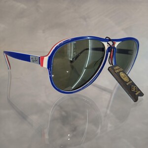 May include: Blue, white, and red aviator sunglasses with green lenses. The sunglasses have a white and red stripe on the top of the frame and a blue stripe on the bottom. The sunglasses are sitting on a white surface.