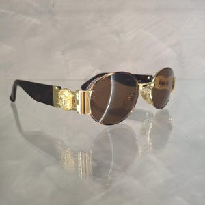 K&ouml;nnte beinhalten: Sonnenbrille mit goldfarbenem Rahmen und braunen Gl&auml;sern. Die Sonnenbrille hat ein goldenes Emblem an den B&uuml;geln und dunkelbraune B&uuml;gel im Schildpattmuster. Die Gl&auml;ser sind oval.