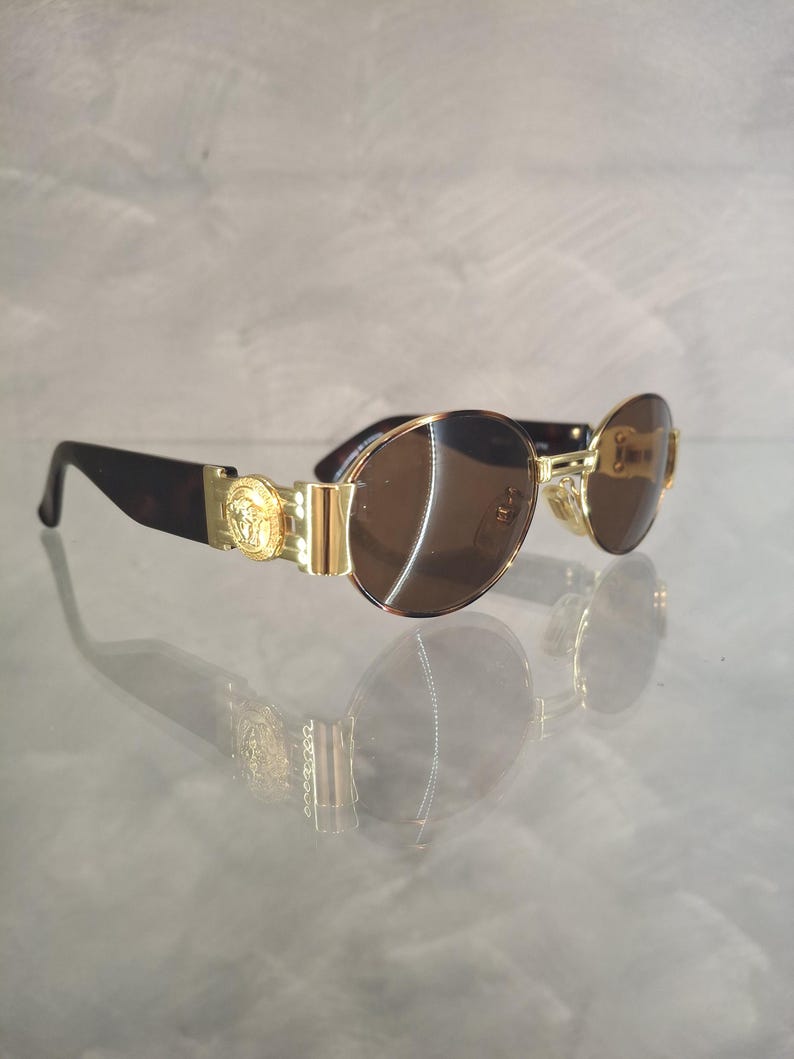 K&ouml;nnte beinhalten: Sonnenbrille mit goldfarbenem Rahmen und braunen Gl&auml;sern. Die Sonnenbrille hat ein goldenes Emblem an den B&uuml;geln und dunkelbraune B&uuml;gel im Schildpattmuster. Die Gl&auml;ser sind oval.