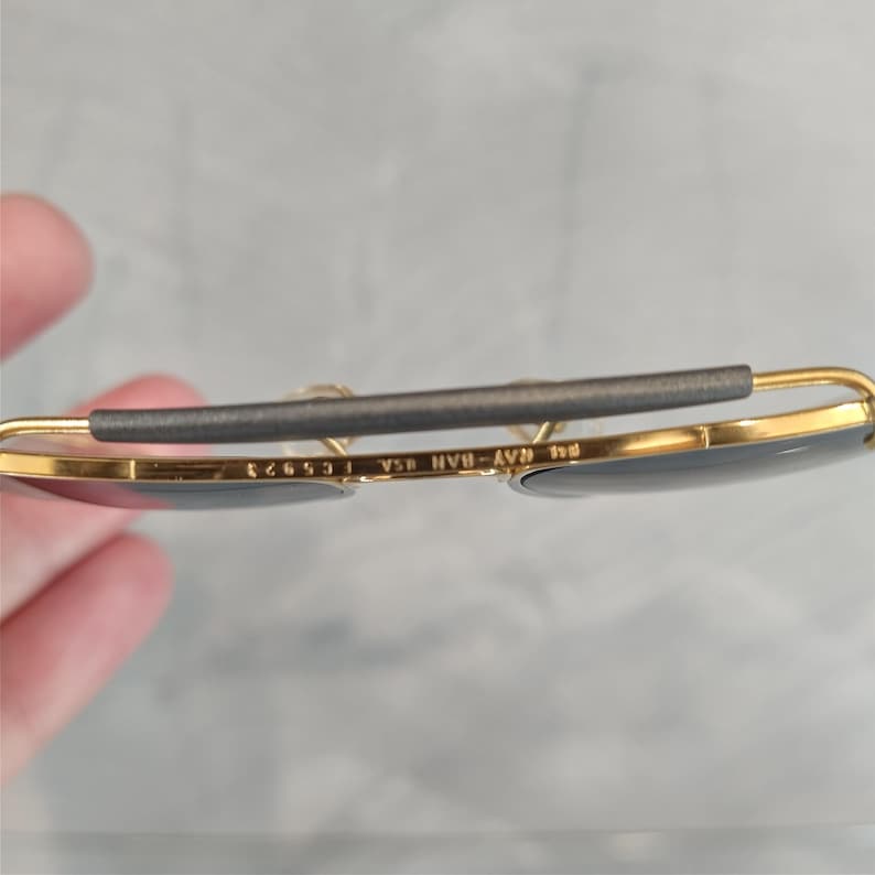 Puede incluir: Gafas de sol aviador doradas con lentes grises. Las gafas tienen una banda de cuero gris con el texto "Ray-Ban B&L USA 12653" grabado en la banda.