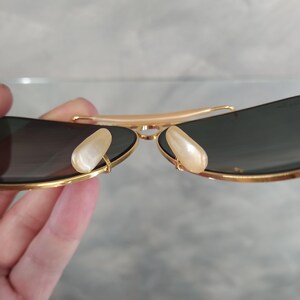 Puede incluir: Gafas de sol aviador doradas con lentes oscuros y almohadillas nasales de perla blanca.