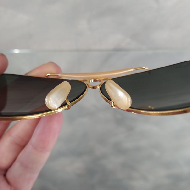 Puede incluir: Gafas de sol aviador doradas con lentes oscuros y almohadillas nasales de perla blanca.