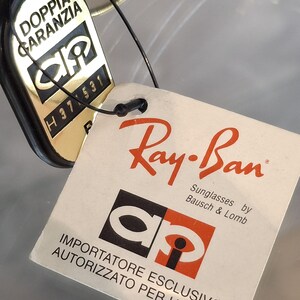 May include: Close-up of Ray-Ban sunglasses tags. One tag is gold and black with the text "DOPPIA GARANZIA" and "H37 531". The other tag is white with the Ray-Ban logo and text "Sunglasses by Bausch & Lomb".