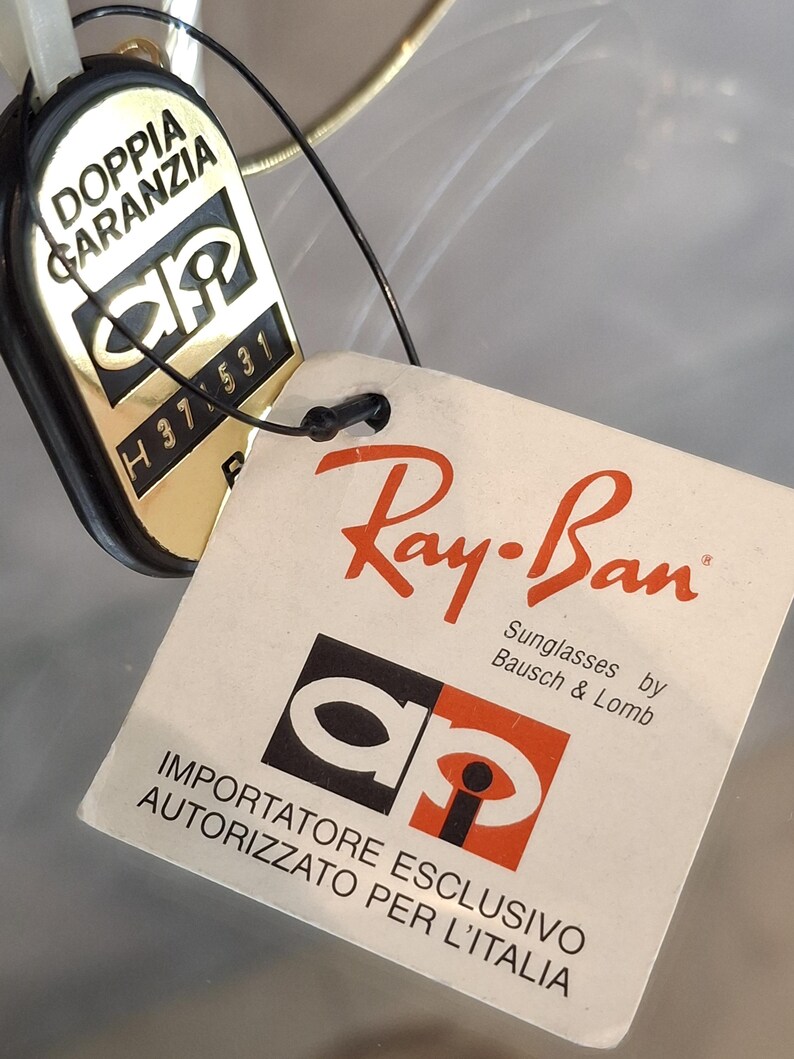 May include: Close-up of Ray-Ban sunglasses tags. One tag is gold and black with the text "DOPPIA GARANZIA" and "H37 531". The other tag is white with the Ray-Ban logo and text "Sunglasses by Bausch & Lomb".
