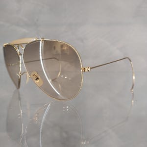 May include: Gold aviator sunglasses with brown gradient lenses. The sunglasses have a double bridge and a thin metal frame.