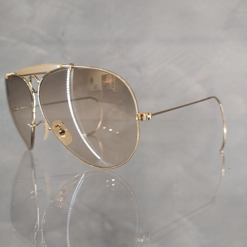 May include: Gold aviator sunglasses with brown gradient lenses. The sunglasses have a double bridge and a thin metal frame.
