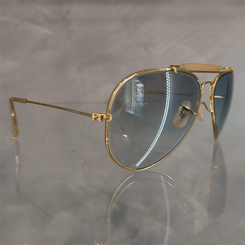 May include: Gold aviator sunglasses with blue gradient lenses. The sunglasses have a metal frame and are reflected in a mirror-like surface.
