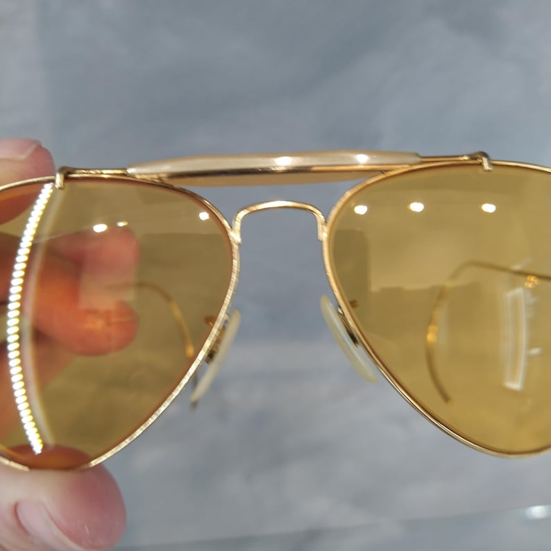 May include: Gold-framed aviator sunglasses with yellow-tinted lenses. The sunglasses have a double bridge and white nose pads. The lenses are teardrop-shaped and reflect light. The overall design is classic and stylish.