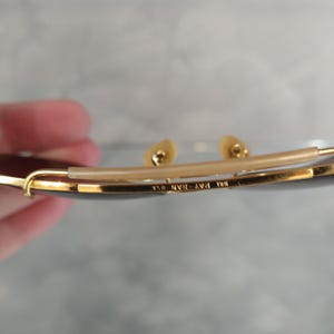 May include: Close-up of the inside of a pair of gold-tone Ray-Ban sunglasses. The sunglasses have a brown lens and the text "Ray-Ban 130 USA" is engraved on the metal frame.