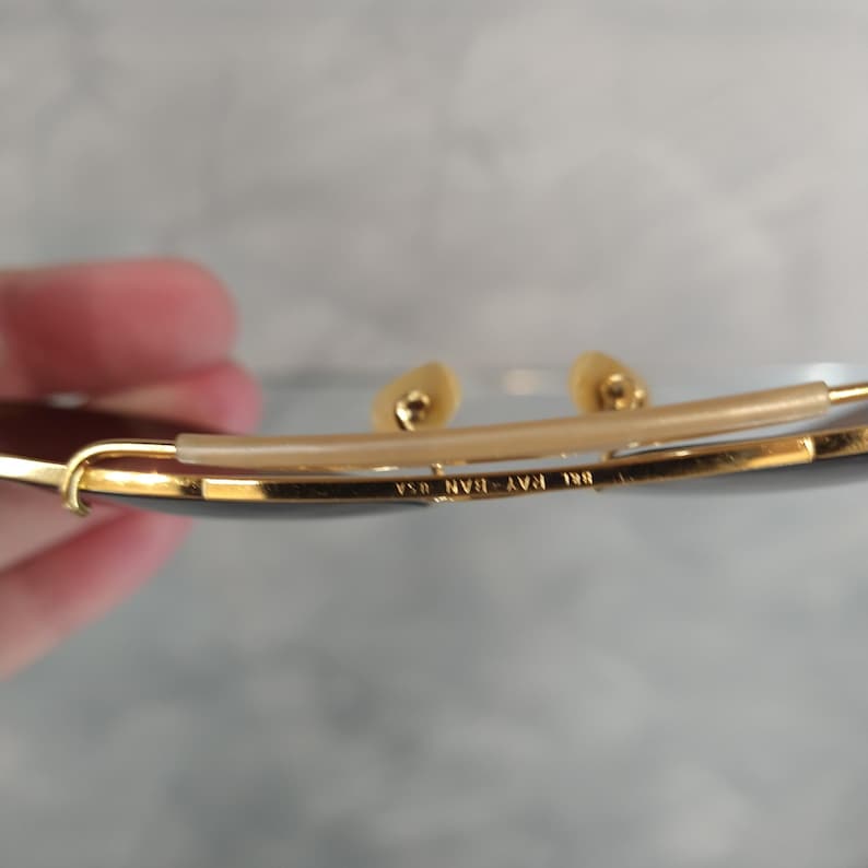 May include: Close-up of the inside of a pair of gold-tone Ray-Ban sunglasses. The sunglasses have a brown lens and the text "Ray-Ban 130 USA" is engraved on the metal frame.